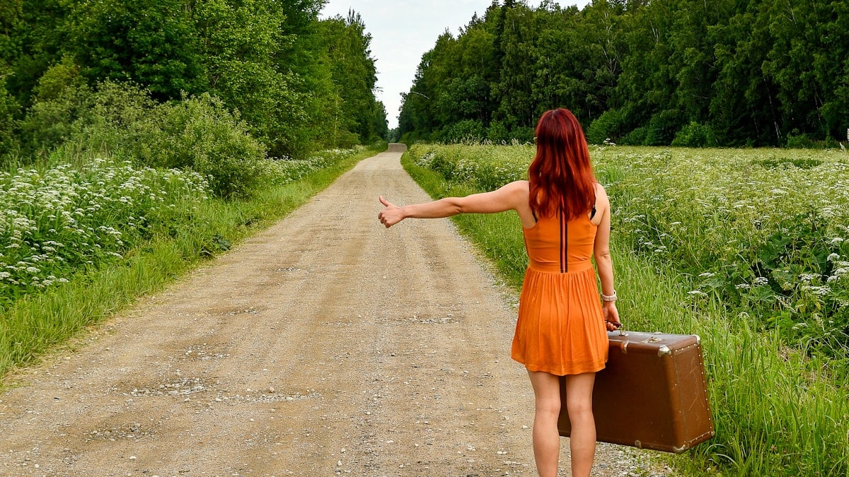 A young red-haired woman in a bright orange dress standing on an empty gravel road with a vintage suitcase, hitchhiking towards a new adventure. 📸 Photo by me IG: @margoevardson